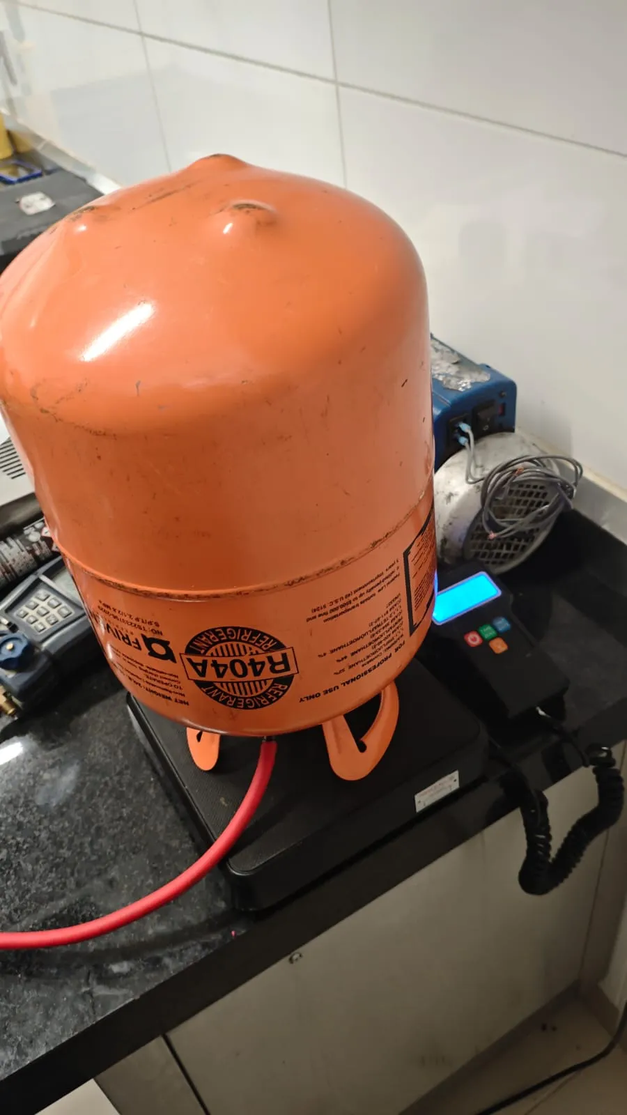 Cilindro laranja de fluido R404A sobre balança eletrônica de carga, ligado por mangueira vermelha, com bomba de vácuo ao fundo